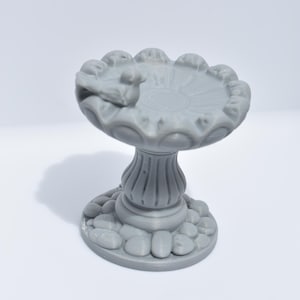 May include: A gray bird bath with a pedestal base and a scalloped rim. The bird bath has a small bird sculpture on the edge of the basin and is surrounded by small stone details at the base.