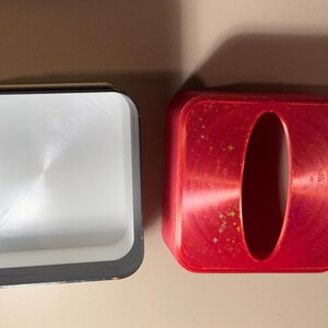 May include: Two square containers. One is open, with a silver interior and black trim. The other is red with an oval opening. Both are on a light brown surface. The red container has a metallic finish with small gold flecks.