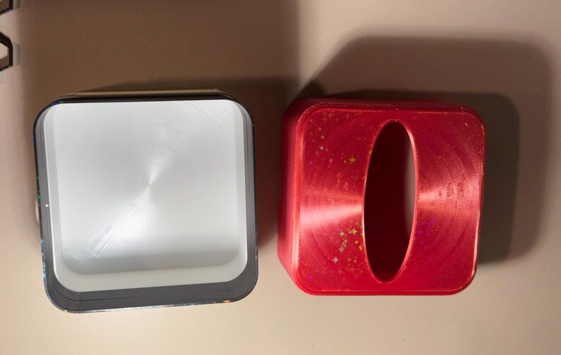 May include: Two square containers. One is open, with a silver interior and black trim. The other is red with an oval opening. Both are on a light brown surface. The red container has a metallic finish with small gold flecks.