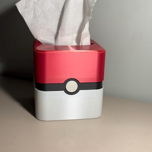 May include: A red, black, and white tissue box with a button detail, resembling a Pok&eacute; Ball. White tissues are visible at the top. The box is on a neutral surface.