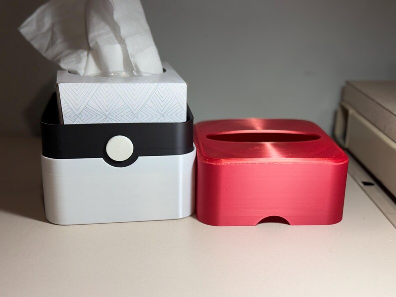 May include: Two tissue box holders. One is white with a black top and a white circular button. The other is a solid red box. The white box contains a tissue box with white tissues. The tissue box has a geometric pattern.