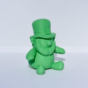 May include: A solid green figurine of a leprechaun, complete with a top hat, beard, and a seated pose. The figure is a single shade of green and sits on a white surface. The hat has a small square detail.