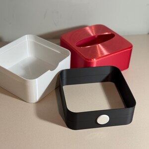May include: Three square containers in white, red, and black. The white container is open, the red has an oval opening, and the black has a beige interior and a white button. The containers are on a light-coloured surface.