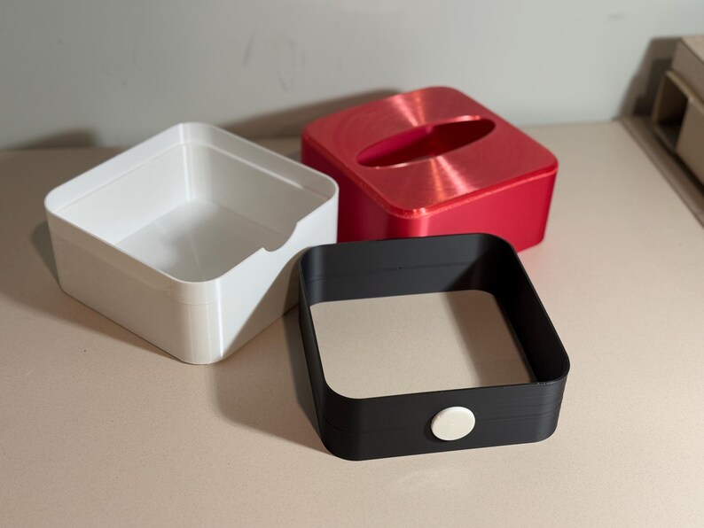 May include: Three square containers in white, red, and black. The white container is open, the red has an oval opening, and the black has a beige interior and a white button. The containers are on a light-coloured surface.