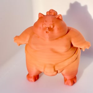 Könnte beinhalten: Eine orangefarbene 3D-gedruckte Figur eines Sumo-Ringers mit katzenartigem Gesicht. Die Figur steht mit ausgestreckten Armen.