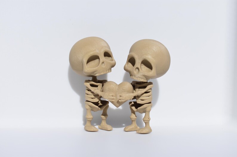May include: Two small skeleton figurines holding a heart-shaped object. The skeletons are beige and have a detailed skeletal structure. The heart is also beige.