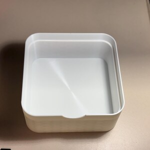 May include: A white, square plastic container with a slightly recessed interior. The container has rounded corners and a small notch on one side. The container is empty and sits on a light-coloured surface.