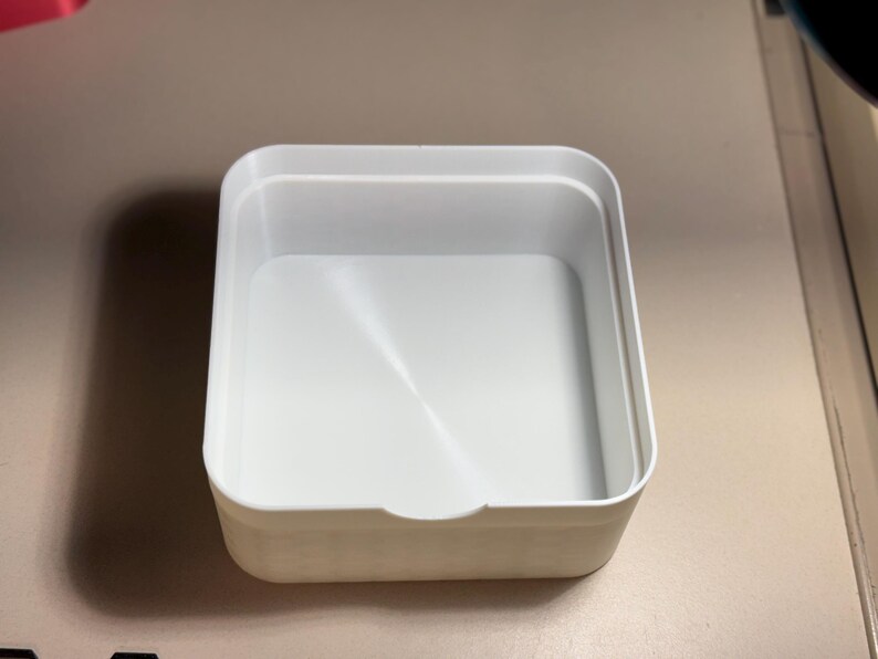 May include: A white, square plastic container with a slightly recessed interior. The container has rounded corners and a small notch on one side. The container is empty and sits on a light-coloured surface.