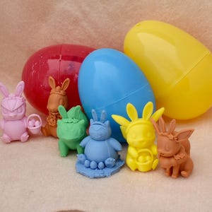 May include: A set of seven colorful Easter egg-shaped figurines, including a pink bunny holding a basket of eggs, a brown bunny with a crown, a green three-headed creature, a blue bunny with a crown, a yellow bunny with a crown, and two brown bunnies with crowns.