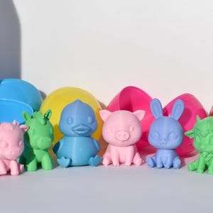 May include: A set of seven colourful plastic animal figurines, including a pink pig, a blue duck, a green unicorn, a pink pig, a blue bunny, a green goat, and a green dragon. The figurines are sitting on a white surface.