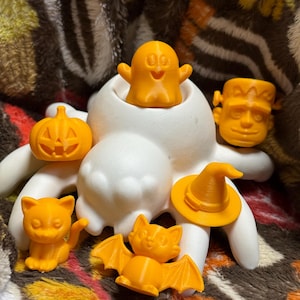 Op de afbeelding: Een verzameling oranje Halloween-thema beeldjes, waaronder een spook, Frankenstein's monster, een pompoen, een kat, een vleermuis en een heksenhoed. De beeldjes zijn gerangschikt rond een witte, abstracte vorm.