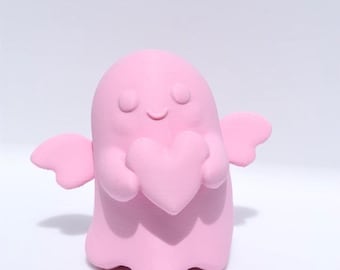 May include: A pink ghost figurine holding a pink heart. The ghost has closed eyes and small wings. The figurine is a solid pink color and is set against a white background.