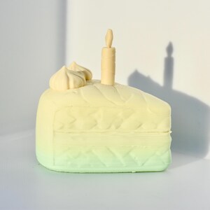 May include: A green and yellow cake slice with white frosting and a single candle on top.