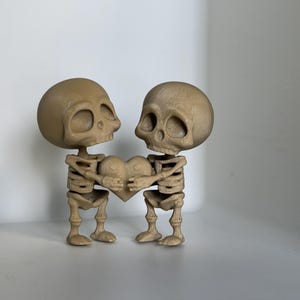 May include: Two small, brown skeleton figurines holding a heart-shaped object. The skeletons are standing upright and facing each other. The heart is brown and has a textured surface.