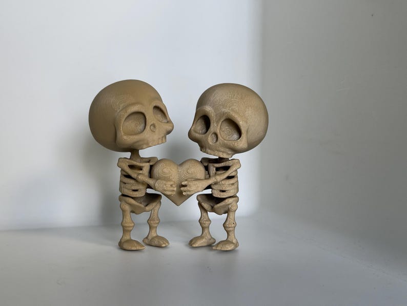 May include: Two small, brown skeleton figurines holding a heart-shaped object. The skeletons are standing upright and facing each other. The heart is brown and has a textured surface.