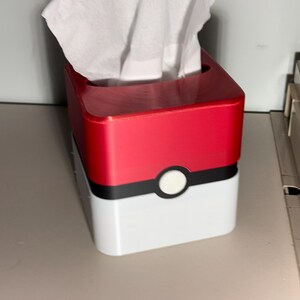 May include: A red and white tissue box, with a black stripe and a white circle, designed to look like a Pokeball. White tissues are visible at the top. The box is on a light grey surface.