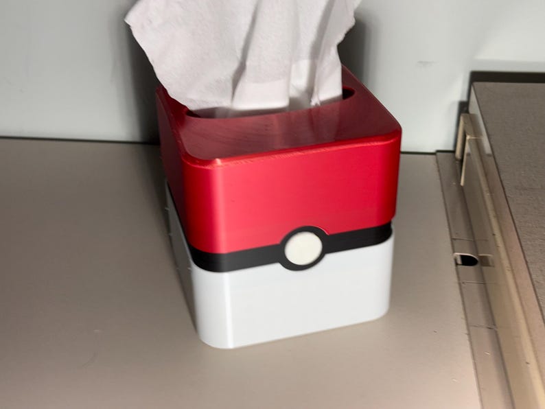 May include: A red and white tissue box, with a black stripe and a white circle, designed to look like a Pokeball. White tissues are visible at the top. The box is on a light grey surface.