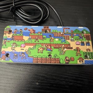 May include: A rectangular video game controller faceplate featuring a colorful pixel art collage of various classic video game characters, including Mario, Link, Mega Man, and Donkey Kong, set against a landscape of green grass and blue water.