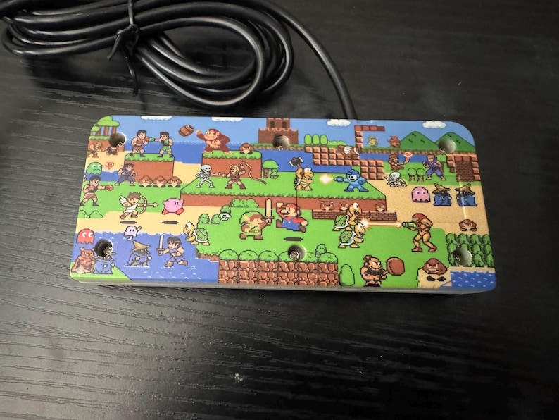 May include: A rectangular video game controller faceplate featuring a colorful pixel art collage of various classic video game characters, including Mario, Link, Mega Man, and Donkey Kong, set against a landscape of green grass and blue water.