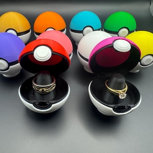 May include: A collection of colorful, spherical ring boxes, resembling Poké Balls, with white bases and various colored tops. Two boxes are open, displaying rings on black stands. The colors include yellow, orange, red, pink, white, blue, and green.