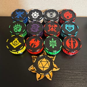 May include: A collection of black and colored game tokens with various symbols, including a leaf, flame, and shield. The tokens have a circular design with a colored edge and a detailed emblem on top. A golden, flower-shaped token is also present.