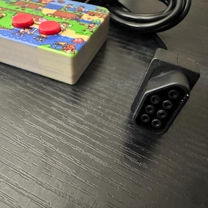 May include: A rectangular video game controller featuring a colorful pixelated landscape graphic on its top surface, two red circular buttons, and a black rectangular button labeled START. A black cable with a nine-pin connector is attached to the device.