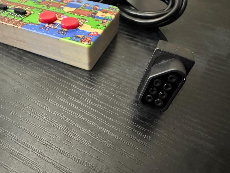 May include: A rectangular video game controller featuring a colorful pixelated landscape graphic on its top surface, two red circular buttons, and a black rectangular button labeled START. A black cable with a nine-pin connector is attached to the device.