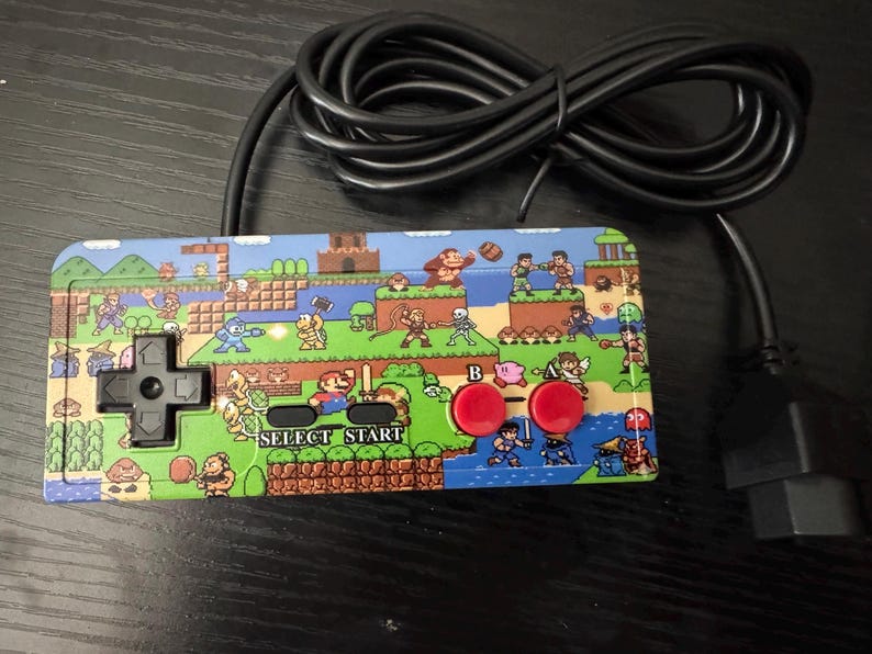 May include: A rectangular video game controller featuring a colorful pixel art graphic of various video game characters. The controller has a black directional pad, two red circular buttons labeled A and B, and black buttons labeled SELECT and START.