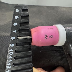 May include: A black ruler with white markings is used to measure the distance of a welding torch tip. The torch has a pink nozzle with the number 8 printed on it. The torch is black and has a white ring.