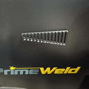 K&ouml;nnte beinhalten: Schwarze Oberfl&auml;che mit dem gelb-wei&szlig;en "PrimeWeld"-Logo. Eine schwarze Metalllehre mit den Zahlen 4 bis 16 befindet sich &uuml;ber dem Logo. Die Lehre hat vertikale Schlitze und dient zum Messen.
