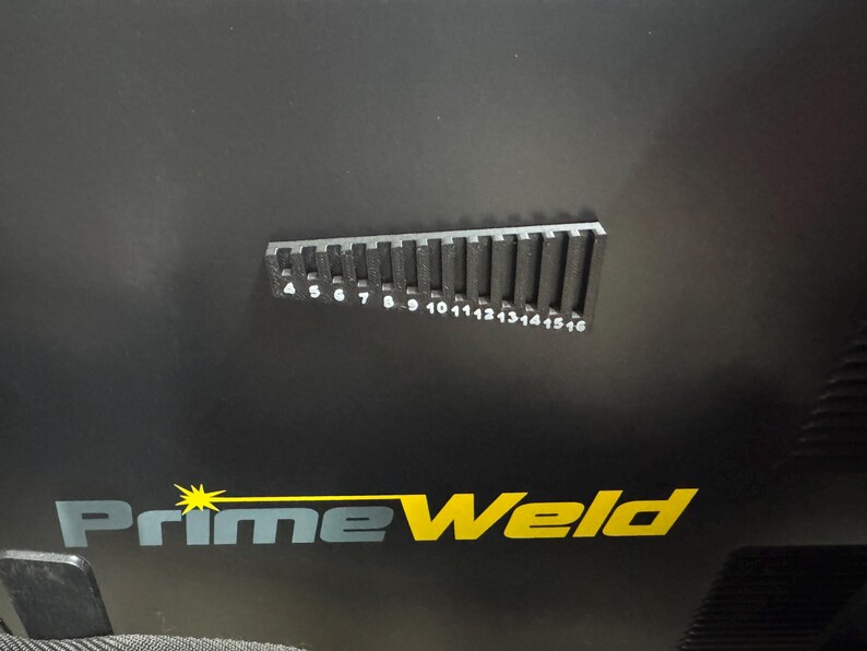 K&ouml;nnte beinhalten: Schwarze Oberfl&auml;che mit dem gelb-wei&szlig;en "PrimeWeld"-Logo. Eine schwarze Metalllehre mit den Zahlen 4 bis 16 befindet sich &uuml;ber dem Logo. Die Lehre hat vertikale Schlitze und dient zum Messen.