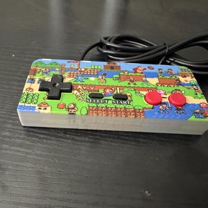 May include: A rectangular video game controller featuring a colorful graphic print of various classic 8-bit video game characters across the top surface. The controller includes a black directional pad, two red circular buttons, and black text labels reading SELECT and START, with a black wired cable attached.