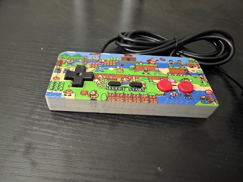 May include: A rectangular video game controller featuring a colorful graphic print of various classic 8-bit video game characters across the top surface. The controller includes a black directional pad, two red circular buttons, and black text labels reading SELECT and START, with a black wired cable attached.