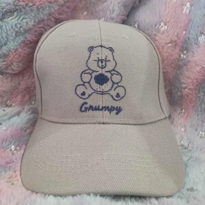 May include: A light gray baseball cap featuring an embroidered blue bear design with a heart on its belly. The word "Grumpy" is embroidered above the brim. The cap is made of a textured fabric.