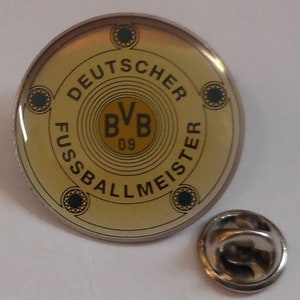 Könnte beinhalten: Eine goldene und schwarze Anstecknadel mit dem Text "DEUTSCHER FUSSBALLMEISTER" um einen Kreis mit dem Text "BVB 09".
