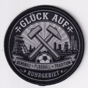 May include: A circular embroidered patch in black and grey featuring crossed mining hammers, a football, and industrial structures. The text reads GLÜCK AUF, BERGBAU FUSSBALL TRADITION, and RUHRGEBIET. The design includes stylized mining towers.