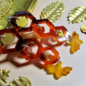 May include: A pair of dangle earrings featuring a honeycomb design with multiple hexagonal shapes in a rich amber color. The earrings are accented with gold-colored metal accents and bee charms.