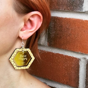 May include: A pair of wooden earrings shaped like a honeycomb with the text "Bee Happy" inside. The earrings are yellow and brown.