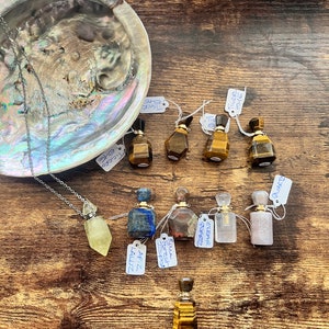 May include: A selection of gemstone perfume bottles and a necklace with a crystal pendant. The bottles are in various colours and shapes, including tiger's eye, lapis lazuli, and clear quartz. The necklace has a yellow crystal pendant. Displayed on a wooden surface.