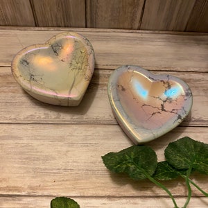 May include: Two iridescent heart-shaped bowls made of white marble with a light pink and blue hue. The bowls are sitting on a light brown wooden surface with green leaves.