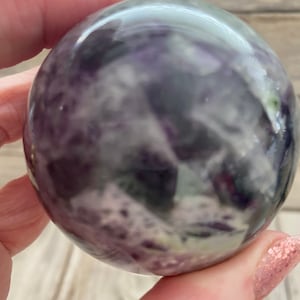 May include: A purple, green, and white fluorite sphere. The sphere is smooth and polished, with a swirling pattern of colors.