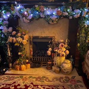 May include: A fireplace mantel decorated with a white floral garland and fairy lights. The fireplace has a black fire grate and is surrounded by a white brick wall. There are candles, lanterns, and other decorative items on the mantel.
