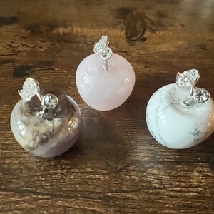 Puede incluir: Tres figuritas de manzana de cristal con acentos de hoja de plata. Las manzanas están hechas de cuarzo rosa, amatista y mármol blanco.