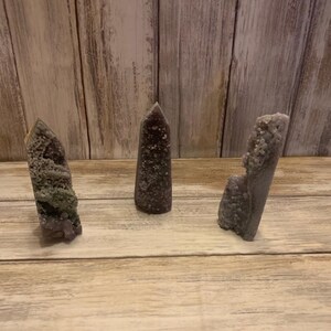 Könnte beinhalten: Drei Amethyst-Kristallspitzen in verschiedenen Formen und Größen. Die Kristalle sind in verschiedenen Violetttönen gehalten, einige haben eine strukturierte, blasenartige Erscheinung. Sie sind auf einer hellen Holzoberfläche angeordnet.