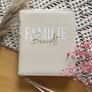 Könnte beinhalten: Eine beige Reißverschlusstasche mit einer weißen Aufschrift "FAMILIE Berndt". Die Tasche hat einen weißen Riemen.
