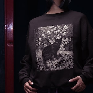 May include: A black sweatshirt featuring a vintage-style print of a black cat sitting in a field of white flowers.