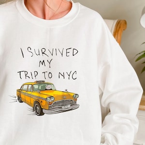 May include: White sweatshirt with the text "I survived my trip to NYC" and a yellow taxi cab illustration.