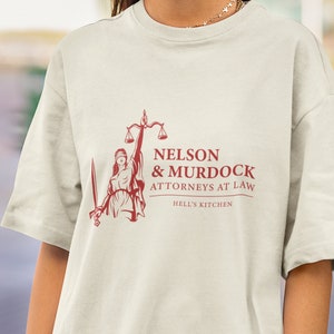 May include: A white t-shirt featuring a red graphic of a blindfolded woman holding scales and a sword. The text "NELSON & MURDOCK ATTORNEYS AT LAW HELL'S KITCHEN" is printed beneath the graphic.