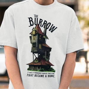Peut inclure: T-shirt blanc avec un graphique d'une maison fantaisiste à plusieurs niveaux avec le texte "The Burrow" au-dessus et "It was a thoroughly magical dwelling that became a home." en dessous.