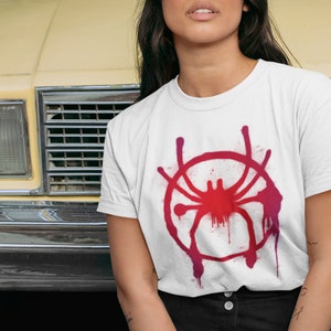 May include: White t-shirt with a red and pink spray paint design of a spider. The spider is in a circle with drips of paint coming from the circle.